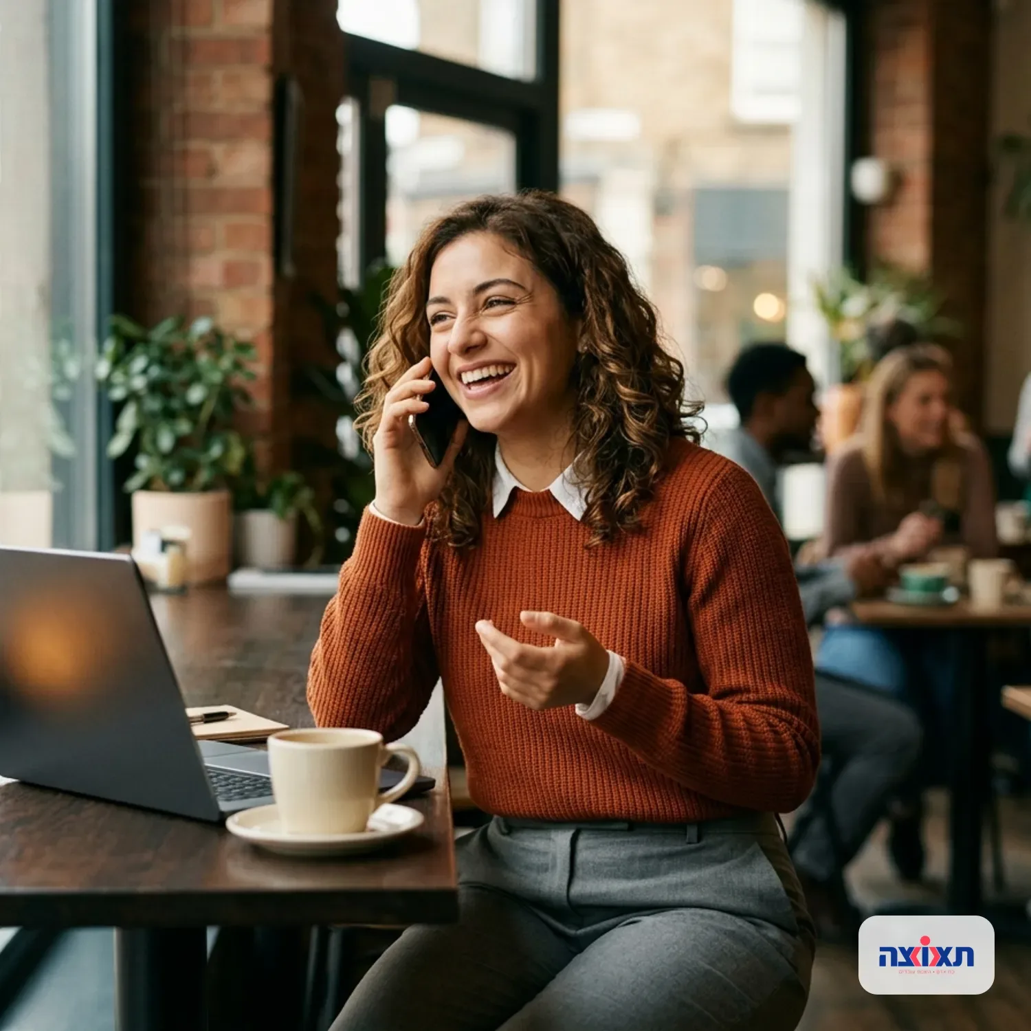 צילום ריאליסטי של אישה לבושה בקז'ואל עסקי (Business Casual) יושבת בבית קפה עם מחשב נייד ומדברת בטלפון בשמחה לאחר קבלת בשורת קבלה לעבודה, אור יום טבעי, פוקוס על הבעת פנים מרוצה