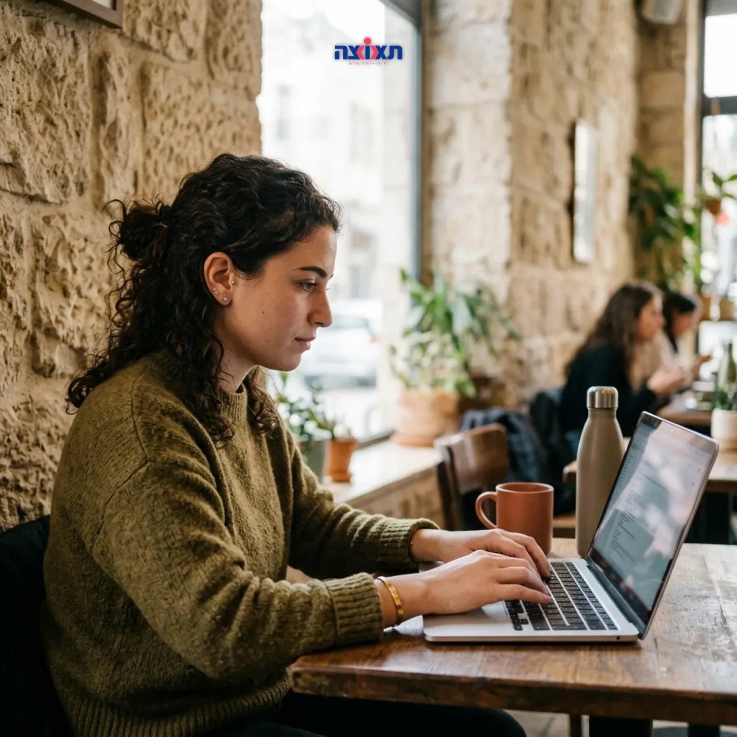 צילום ריאליסטי של סטודנטית עובדת עם מחשב נייד בבית קפה ירושלמי מואר, ברקע קיר אבן ירושלמית, תאורת יום טבעית