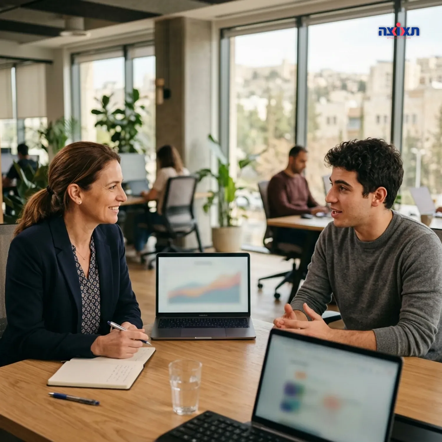 צילום מציאותי של פגישת עבודה במשרד מודרני בהר חוצבים, מנהלת כוח אדם מקשיבה לעובד צעיר, פוקוס חד ותאורה טבעית חמימה